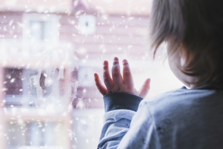 A young child, seen from behind, presses a hand against a windowpane, looking out at falling snow