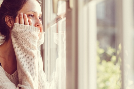A pensive woman in a light sweater looks out a window, hands on her face