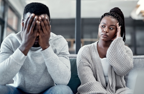 A distressed man covers his face, while a woman with a pensive expression sits beside him