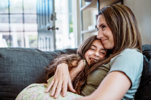 A young girl cuddles with her mother on the living room sofa at home. Visual concept for a blog discussing how a co-parenting app centralizes communication, keeping the child out of parental conflict.