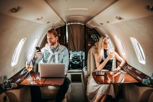 Wealthy man looking at his cell phone with a laptop in front of him facing away from woman looking at her cell phone in airplane